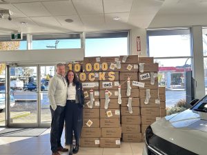 Brad and Bethan Ostermann standing inside Kia Victoria beside a wall of boxes labeled “10,000 Socks,” representing the dealership’s annual fundraiser with Cool Aid Society to provide warm socks and essentials for people in need in Victoria, BC.
