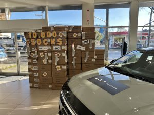 Kia Victoria dealership showroom featuring new Kia vehicles and a large display of stacked boxes labeled “10,000 Socks,” part of the dealership’s annual fundraising campaign with Cool Aid Society to support people in need in Victoria, BC.
