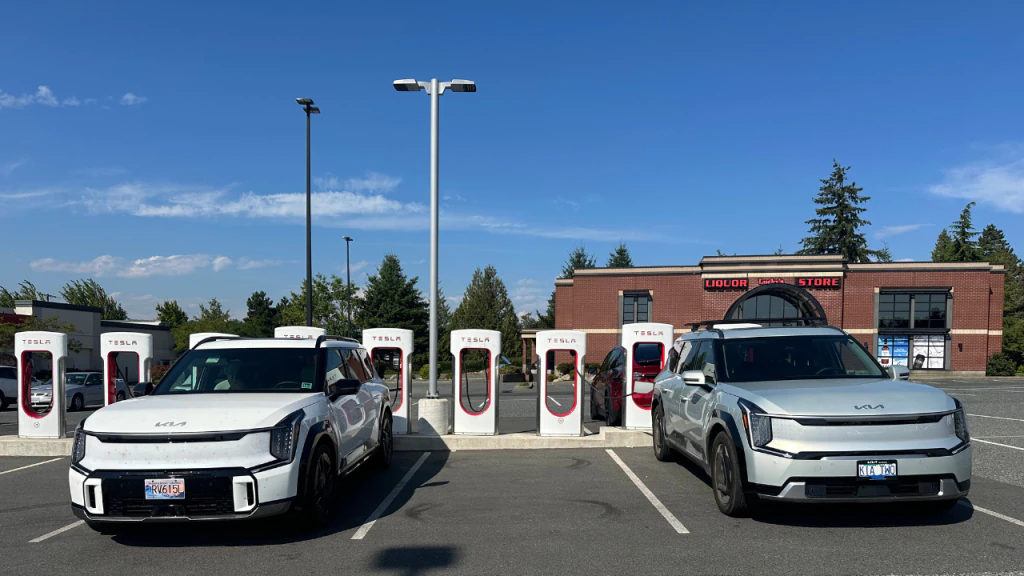Two Kia EV9 SUVs parked at a charging station on Vancouver Island with Tesla Superchargers in the background.